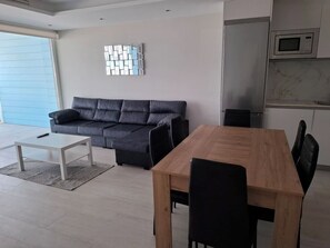 A modern living room with a sofa, a coffee table, a dining table with chairs, and a kitchen area with a microwave. - Luxury Marina Resort | Rock Views & Pools - Apartment (Gibraltar)