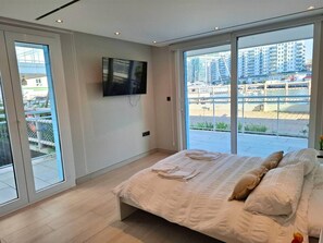 Ein modernes Schlafzimmer mit einem großen Bett, einem flachbildfernseher an der Wand und einem Balkon mit Blick auf hohe Gebäude.