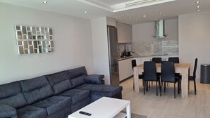 A modern living room with a sectional sofa, a white coffee table, and a dining area with a wooden table and chairs. - Luxury Marina Resort | Rock Views & Pools - Apartment (Gibraltar)