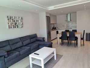 A modern living room with a sectional sofa, a white coffee table, and a dining area with a wooden table and chairs. - Luxury Marina Resort | Rock Views & Pools - Apartment (Gibraltar)