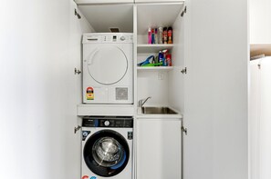 A compact laundry room with a front-loading washer and dryer, a small sink, and shelves with cleaning supplies. - Lake View 1-bed Apartment Canberra Centre (Canberra)