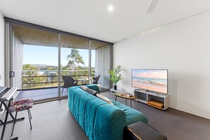 A modern living room with a sofa, a piano, a TV, and a balcony with a view. - Lake View 1-bed Apartment Canberra Centre (Canberra)