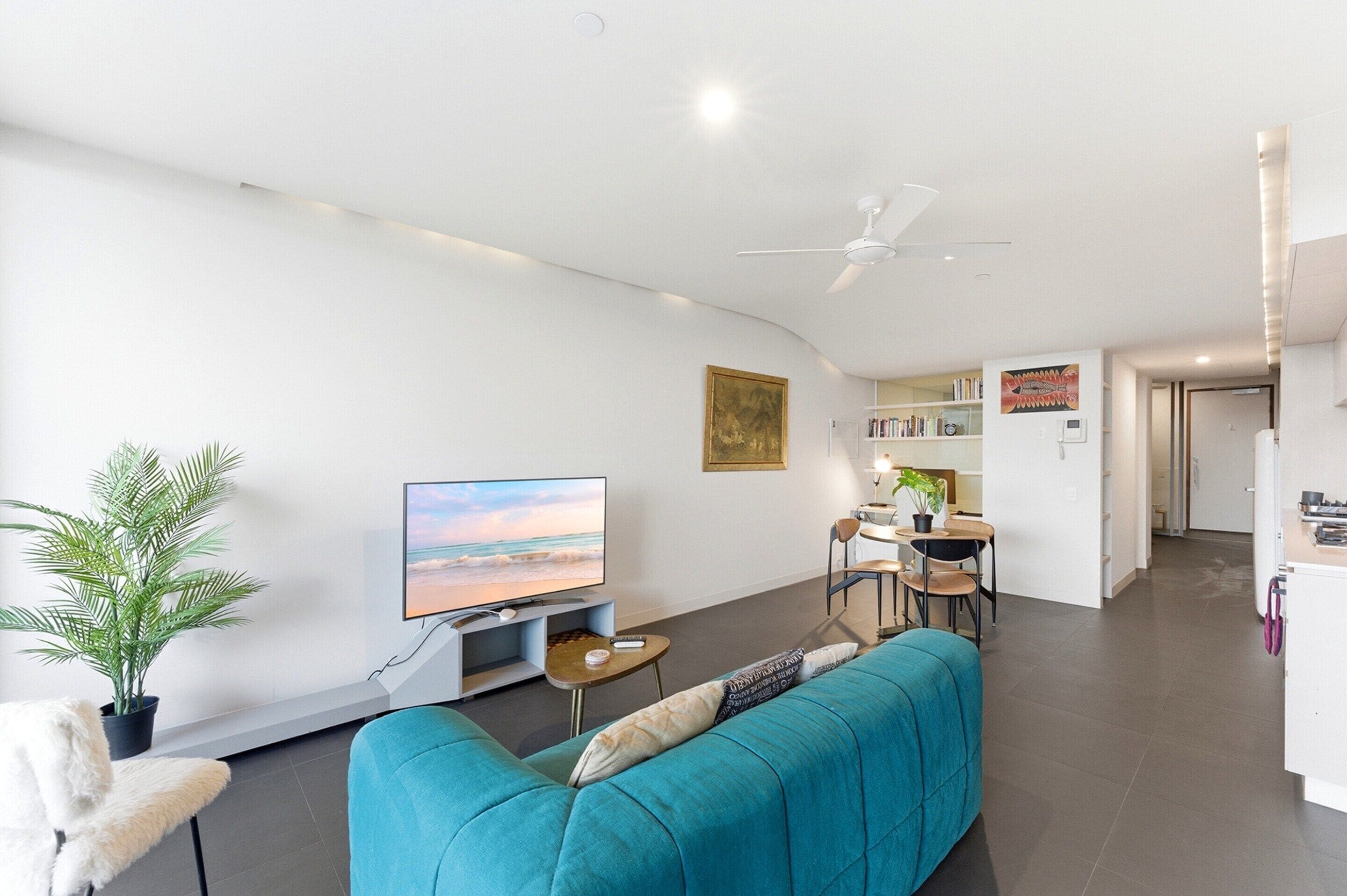 A modern living room with a flat-screen TV, a teal sofa, a dining area with a table and chairs, and a kitchen in the background.