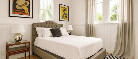A bedroom with a large bed, two bedside tables with lamps, a striped rug, and two posters on the wall.