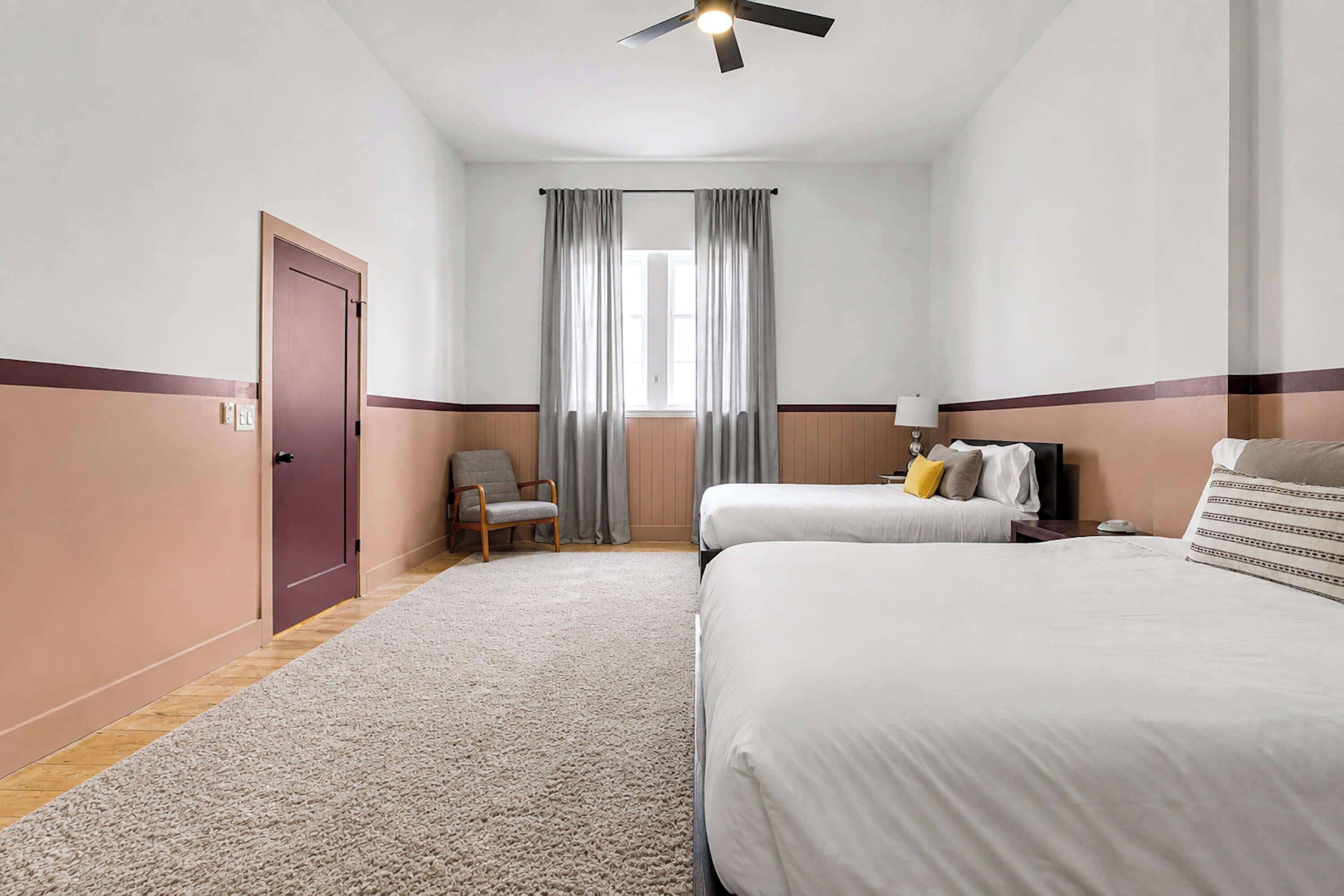 A hotel room with two beds, a chair, a desk, and a ceiling fan.