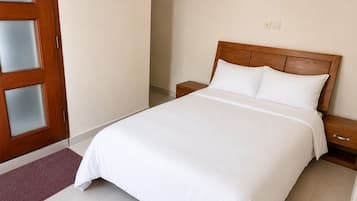 Een slaapkamer met een houten hoofdeinde, een witte spreidekbed, een houten nachtkastje en een paarse deurmat.