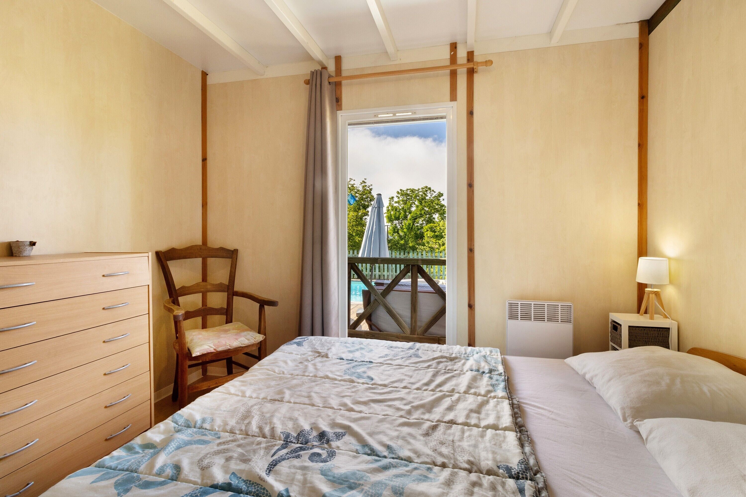 Une chambre à coucher comprenant un lit, une chaise en bois, une commode, une table de chevet avec une lampe, et une vue sur une piscine et des arbres, visible à travers une porte.