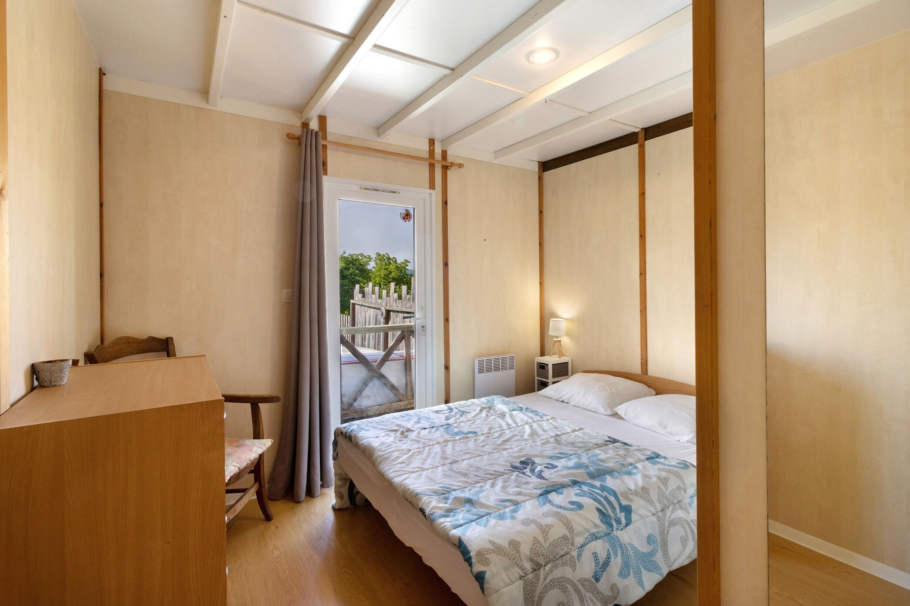 Een slaapkamer met een bed, een bureau, een stoel, een radiator en een raam met uitzicht op groen.