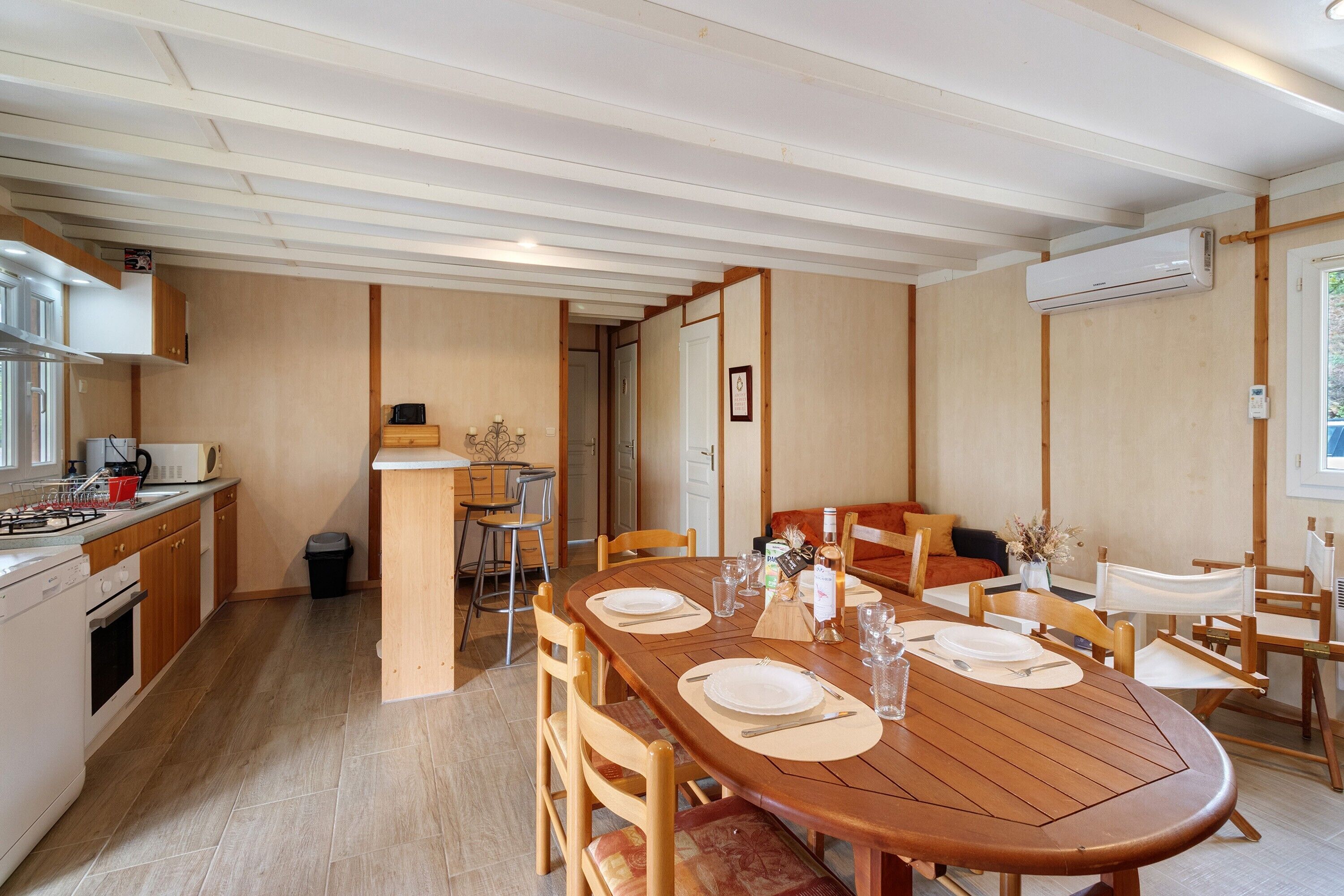 Uma sala de jantar com mesa de madeira posta para quatro, cozinha com eletrodomésticos modernos e vista para o exterior.