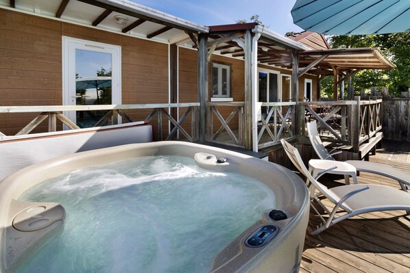A hot tub on a wooden deck with chairs, a wooden fence, and a building with windows.