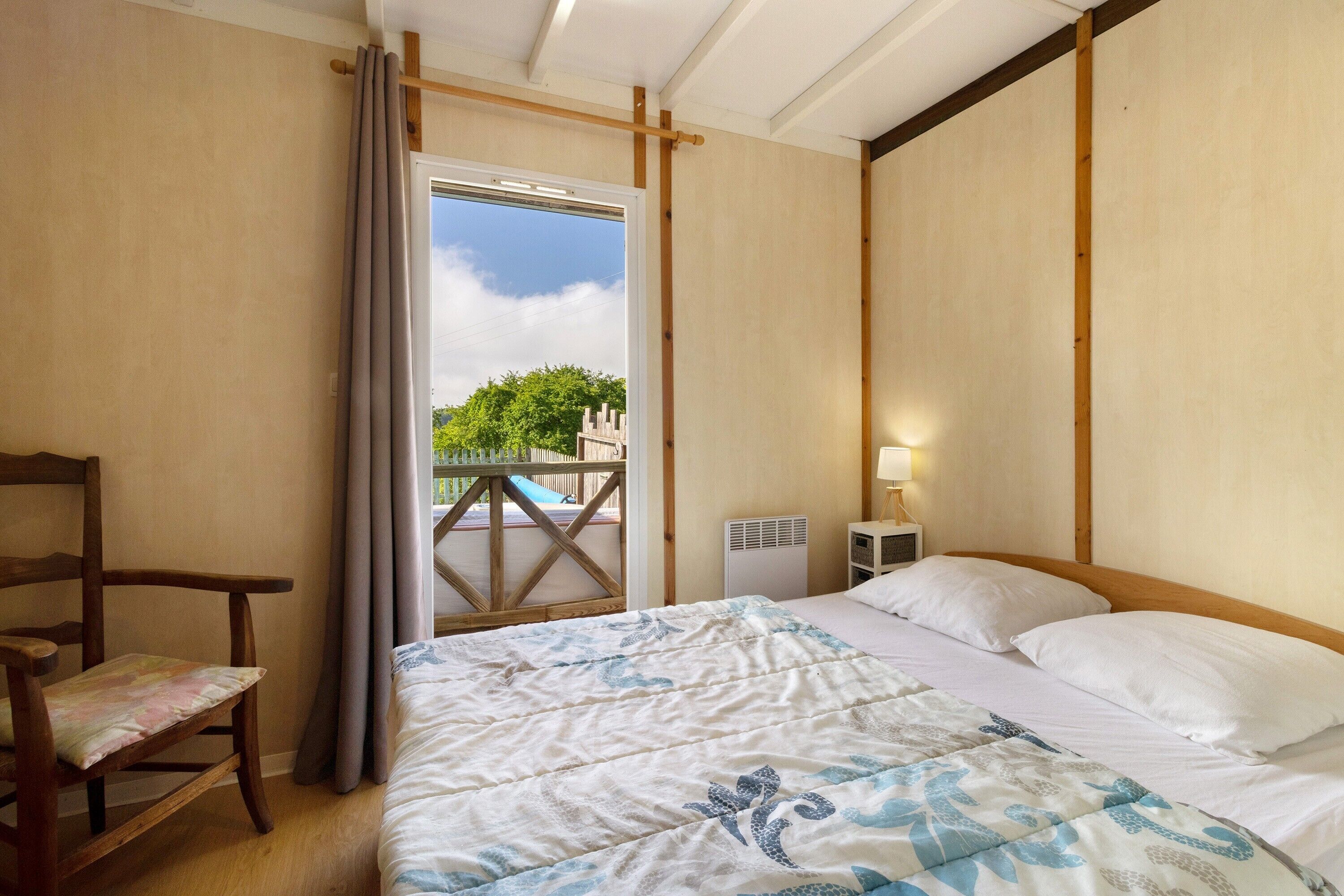 Ein Zimmer mit einem Bett, einem Sessel, einem fenster mit Vorhängen, einer Heizung und Blick ins Grüne.