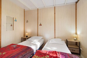 A room with two single beds, wooden walls, a small bedside table with a lamp, and a mirror on the left wall.