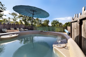 Un bain à remous avec des chaises longues et un parasol, entouré d’une clôture en bois et d’arbres.
