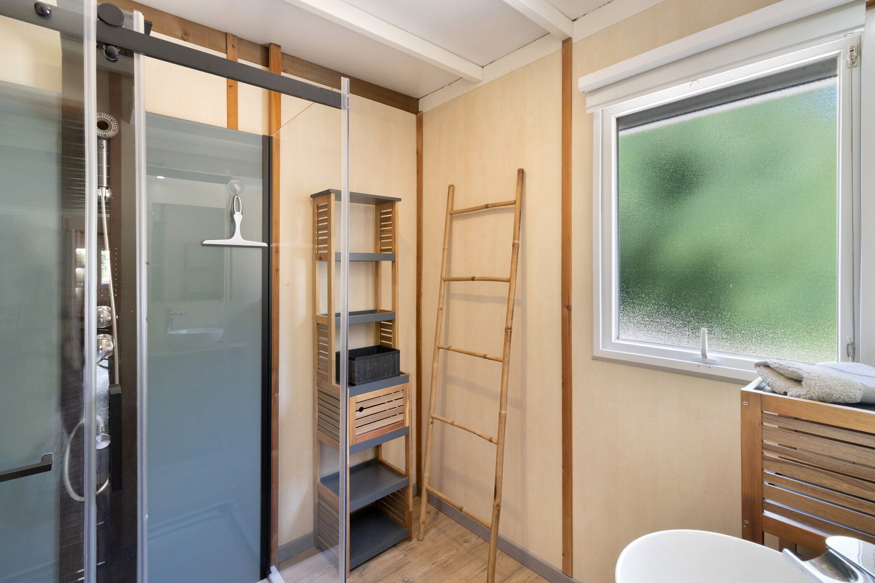 Een badkamer met een douchecabine van glas, een houten wastafelmeubel met een handdoek, en een raam met uitzicht op groen.