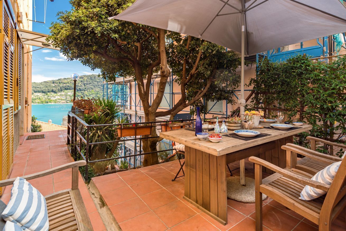 Un espacio para comer al aire libre con mesa y sillas de madera, un paraguas grande y vista al mar.