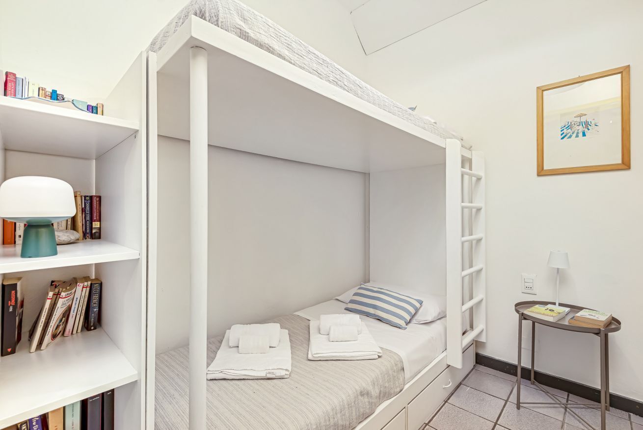 Una cama litera con estructura blanca, una mesita de noche con lámpara y una estantería llena de libros.