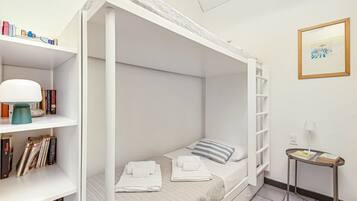 Una cama litera con estructura blanca, una mesita de noche con lámpara y una estantería llena de libros.