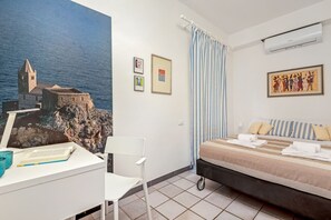 Une chambre d’hôtel comprenant un lit, un bureau avec une chaise, un tableau représentant un paysage côtier, une fenêtre avec des rideaux et un climatiseur fixé au mur.