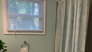 Ein Badezimmer mit einem weißen Waschbecken, einem Handtuchhalter mit Handtuch und einem Fenster mit Jalousien sowie einem Duschvorhang mit geometrischem Muster.