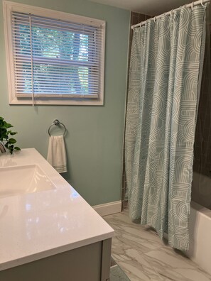 Ein Badezimmer mit einem weißen Waschbecken, einem Handtuchhalter mit Handtuch und einem Fenster mit Jalousien sowie einem Duschvorhang mit geometrischem Muster.