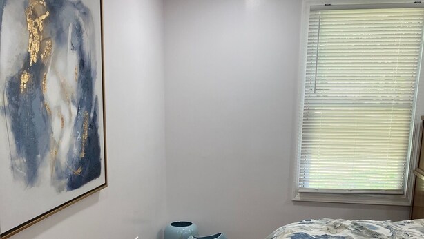 Ein Schlafzimmer mit einem Bett, einem Gemälde, zwei blauen Vasen und einem Fenster mit Jalousien.