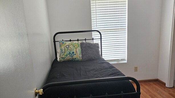 Una cama individual con estructura metálica, una ventana con persianas y suelo de madera.