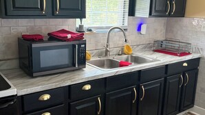 Una cocina con gabinetes oscuros, microondas, lavamanos y ventana con persianas.