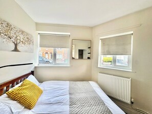 Ein Schlafzimmer mit einem Bett, zwei Fenstern mit Jalousien, einem Heizkörper und einem Spiegel an der Wand.