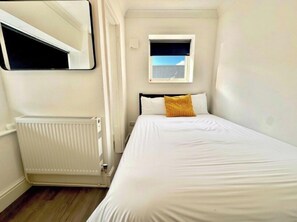Ein kleines Zimmer mit einem Einzelbett, einem Heizkörper, einem Fenster und einem Spiegel an der Wand.