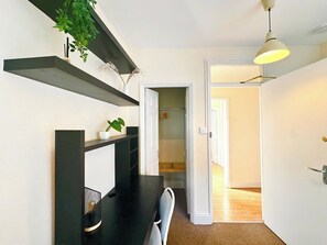 Ein Flur mit Schreibtisch, Stuhl und einer hängenden Pflanze. Eine Tür führt in einen anderen Raum.