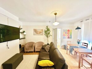 Ein Wohnzimmer mit einer Couch, zwei Sesseln, einem Esstisch mit Stühlen, einem an der Wand montierten Flachbildfernseher und einer Deckenleuchte.