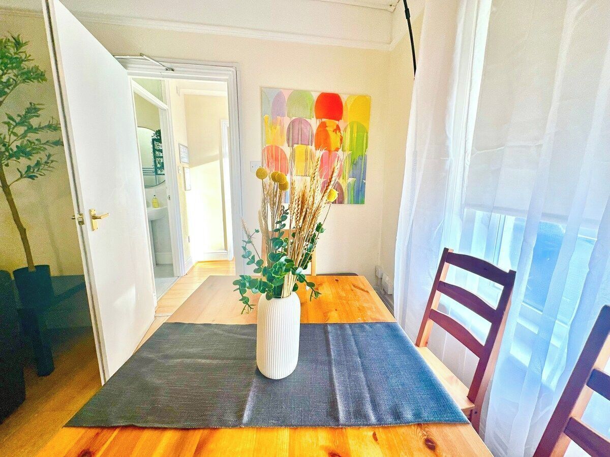 A dining area with a wooden table, a vase with dried plants, and a colorful abstract painting on the wall.
