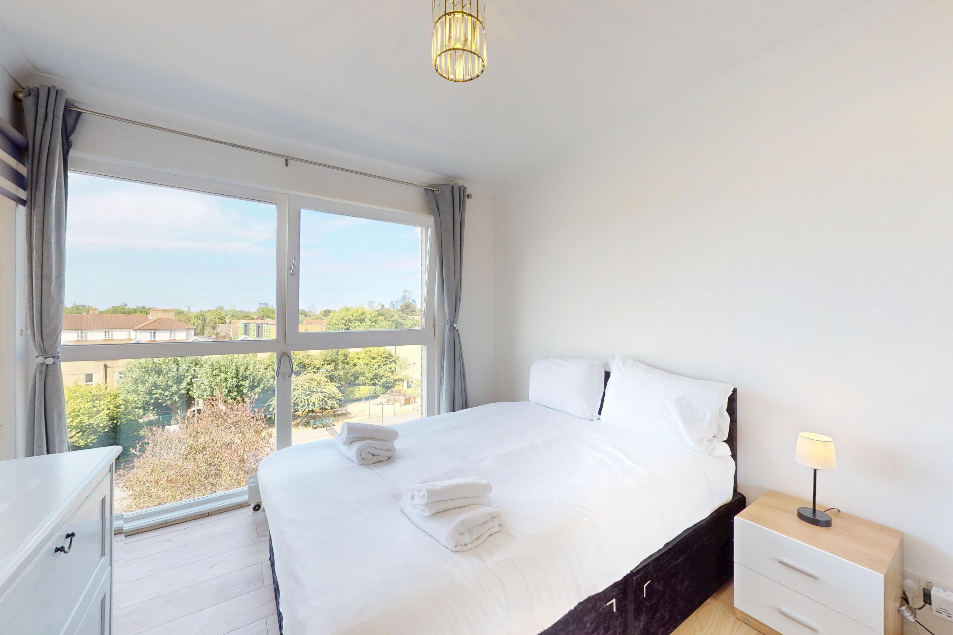 A hotel room with a large window, a bed with white linens, a bedside table with a lamp, and a view of the cityscape.
