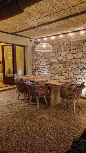 Ein Essbereich mit Steinswand, Holztisch, Korbstühlen und einer hängenden Leuchte.