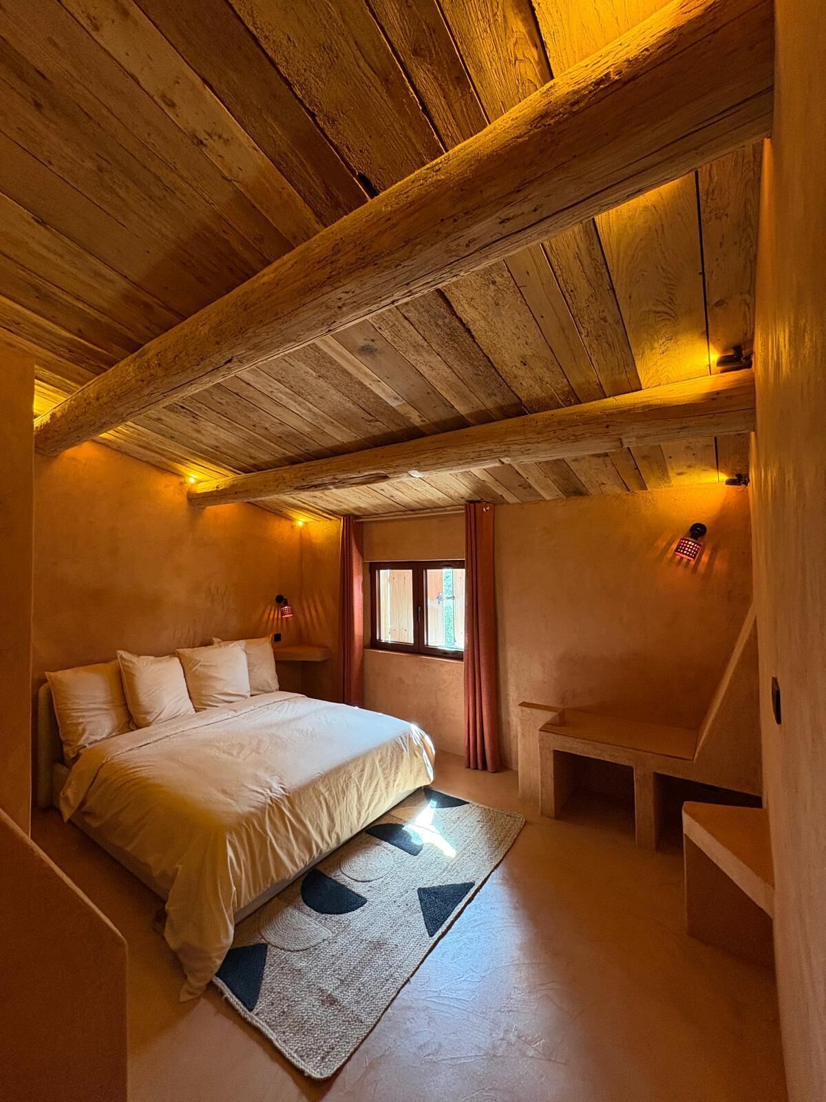 Une chambre avec un plafond en bois, un lit avec une literie blanche, une petite fenêtre avec des rideaux et une table de chevet avec une lampe.