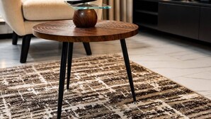 Une table ronde en bois, surmontée d’un plateau en verre, et un vase, le tout posé sur un tapis à motifs.