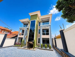 Un edificio moderno de varios pisos con fachada de vidrio central, rodeado por un patio empedrado con plantas.