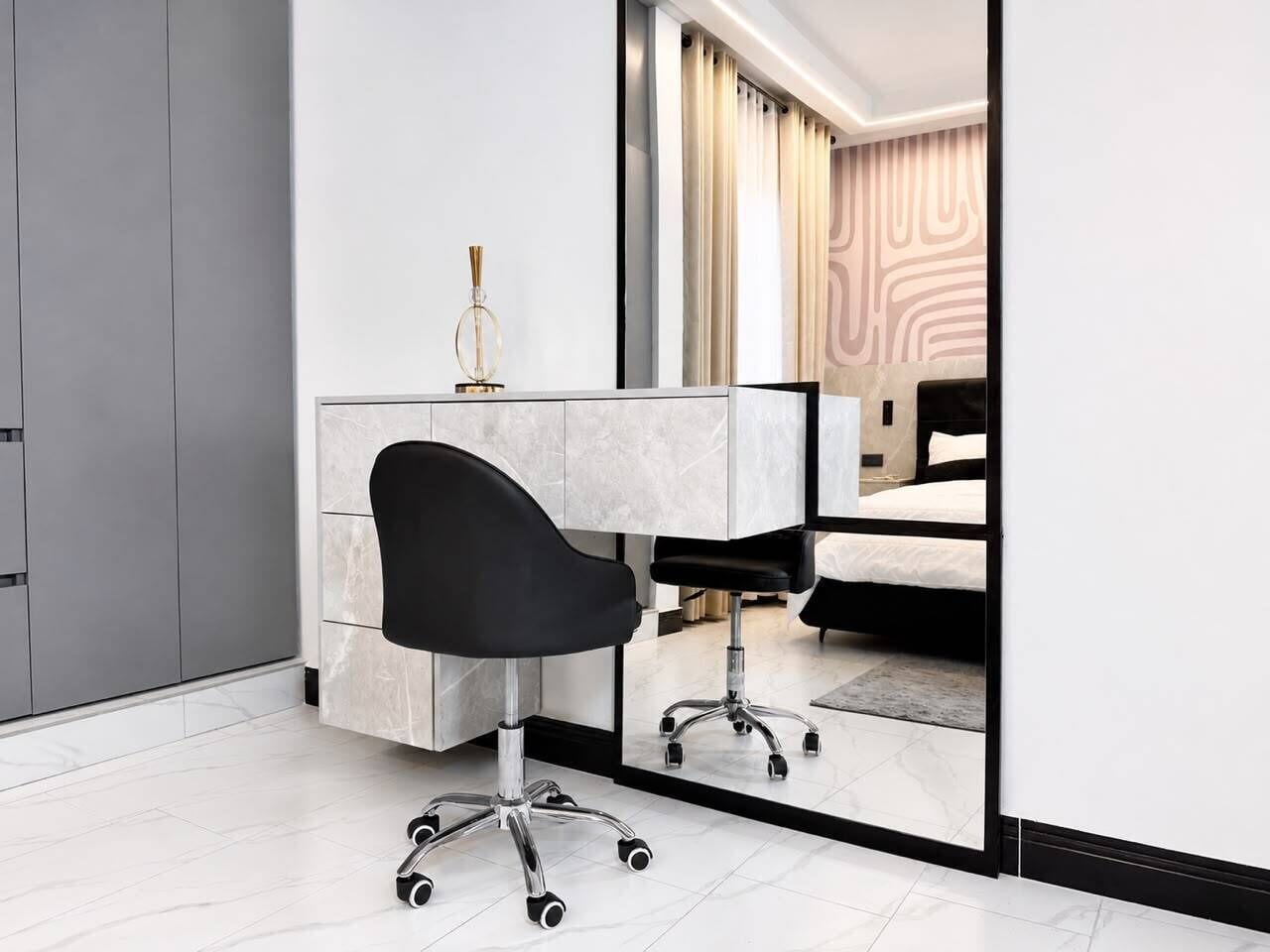 A modern office chair with wheels, a white desk, and a mirror reflecting a bed and curtains.