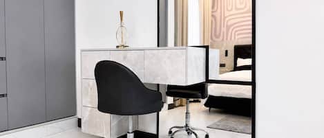 A modern office chair with wheels, a white desk, and a mirror reflecting a bed and curtains.