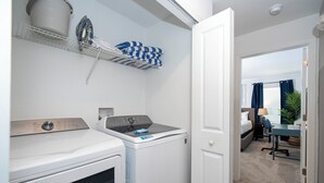 A compact laundry area with a front-loading washer and dryer, a drying rack with folded towels, and a glimpse of a bedroom with a bed and a desk. - Relaxing 4BR ChampionsGate Getaway - Private Pool, BBQ & Family Comfort (Davenport)
