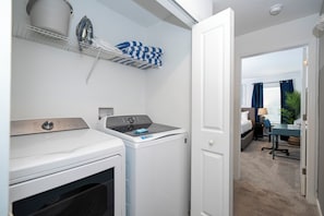 A compact laundry area with a front-loading washer and dryer, a drying rack with folded towels, and a glimpse of a bedroom with a bed and a desk. - Relaxing 4BR ChampionsGate Getaway - Private Pool, BBQ & Family Comfort (Davenport)