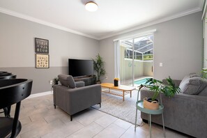 A modern living room with a flat-screen TV, grey sofas, a coffee table, and a sliding glass door leading to a pool area. - ☆☆☆☆☆Price Drop! OCT-NOV - 4BR ChampionsGate - Pool - Themed Rooms! 
☆☆☆☆☆ (Davenport)