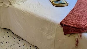 Una cama con ropa de cama blanca, una almohada amarilla y una manta roja. Se ve un ventanal con vistas a la vegetación.