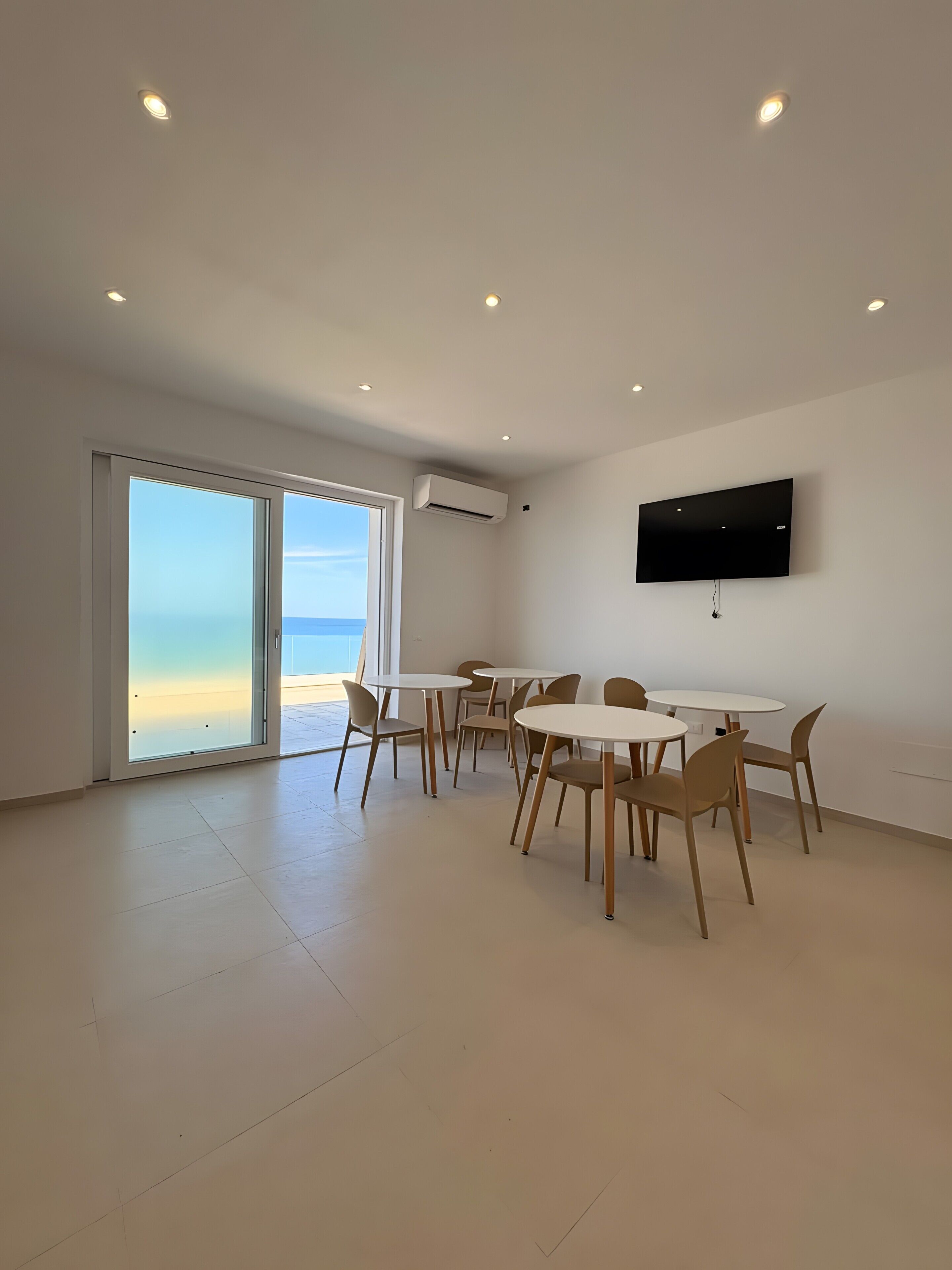 Een ruime kamer met een grote schuifglazen deur naar een balkon, een wandgemonteerde flatscreen-tv en een eethoek met ronde tafels en stoelen.