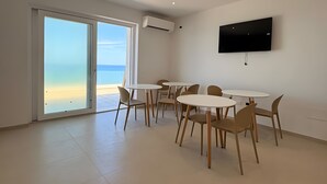 Een ruime kamer met een grote schuifglazen deur naar een balkon, een wandgemonteerde flatscreen-tv en een eethoek met ronde tafels en stoelen.