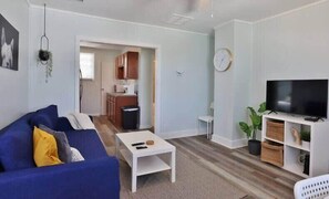 A living room with a blue sofa, a coffee table, a TV on a white stand, and a kitchen in the background. - Cozy Beach Hideaway - 1 block to the beach (Jacksonville Beach)