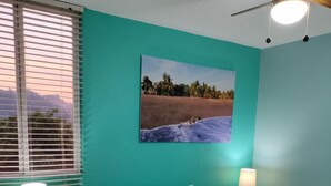 Quarto com cama, duas mesas de cabeceira, janela com persianas, ventoinha no teto e um quadro na parede.