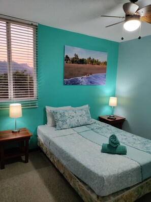 Ein Schlafzimmer mit einem Bett, zwei Nachttischen, einem Fenster mit Jalousien, einem Deckenventilator und einem Wandbild.