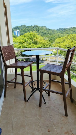 Ein Balkon mit zwei Holzstühlen und einem kleinen runden Tisch, mit Blick auf eine grüne Landschaft mit Bäumen und einem Gebäude in der Ferne.