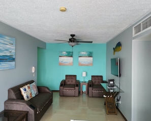 A living room with a teal wall, two reclining chairs, a sofa, a ceiling fan, a TV, and artwork. - Luxury 3BR Jaco Retreat • Pool • Beach • Concierge Included (Jacó)